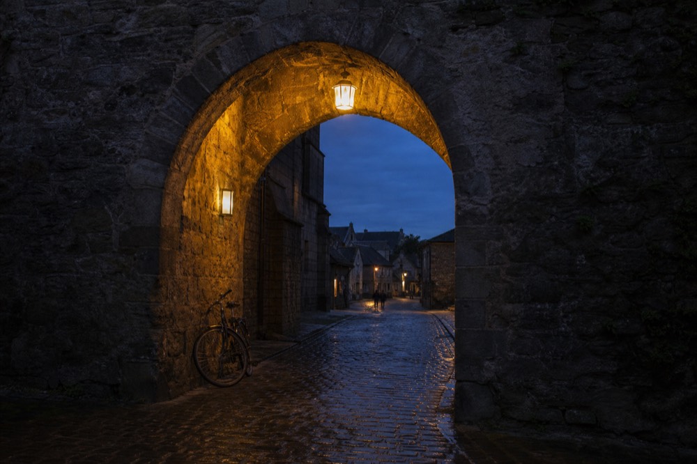 Medieval stone gateway into Saint-Emilion village at dusk with warm lamplight, no text, no logos, no people